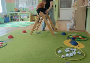 Na zielonym dywanie leży ścieżka sensoryczna. Pani pomaga chłopcu przejść przez drabinkę.