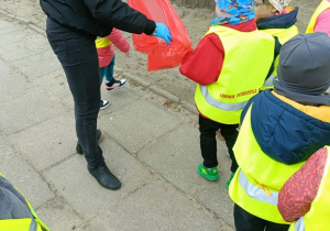Pani trzyma pomarańczowy worek. Dzieci wokół niej szukają śmieci.