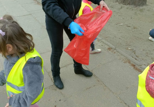 Pani trzyma pomarańczowy worek. Dzieci wokół niej szukają śmieci.