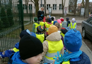 Dzieci w zielonych kamizelkach idą chodnikiem. Po prawej stronie widać kawałek auta.