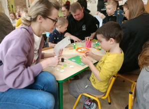 Dzieci siedzą z rodzicami przy stolikach i wykonują kartki.