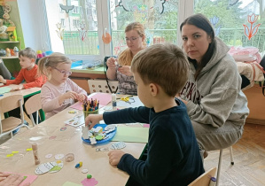 Dwójka dzieci (chłopiec i dziewczynka) siedzą przy stoliku i wykonują z mami kartki.