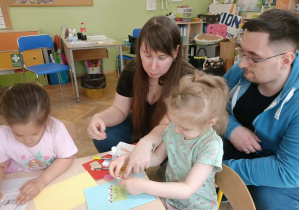Dziewczynka siedzi przy stoliku i wykonuje kartkę z rodzicami. Obok siedzi dziewczynka z ciemnymi włosami.