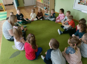 Na zielonym dywanie siedzą w kole dzieci razem z Panią, która czyta im książkę.