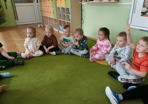 Dzieci siedzą na dywanie i słuchają czytanej książki. Chłopiec w pomarańczowej koszulce zgłasza się do odpowiedzi.