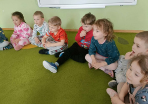 Dzieci siedzą na dywanie i słuchają czytanej książki.