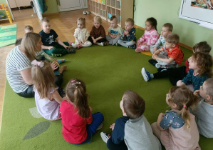 Na zielonym dywanie siedzą w kole dzieci razem z Panią, która czyta im książkę.