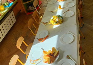 Stół nakryty białym obrusem. Wokół stoją krzesełka.