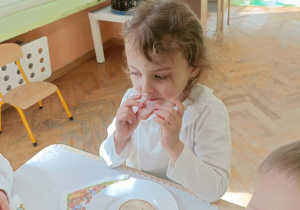 Dziewczynka w białej bluzce siedzi przy stole i je kiełbaskę.