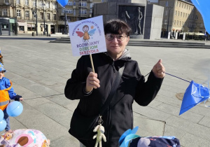 Pani w czarnej kurtce trzyma transparent z logo przedszkola.