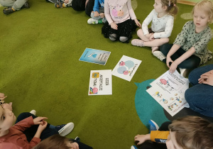 Na zielonym dywanie leżą kolorowe karty. Wokół siedzą dzieci.