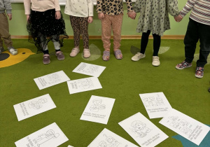 Dzieci stoją i trzymają się za ręce. Na dywanie leżą kolorowanki.