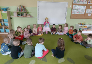 Grupa dzieci siedzi w kole na zielonym dywanie.