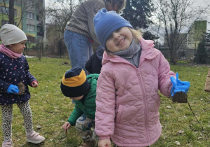 Dziewczynka w różowej kurtce uśmiecha się do zdjęcia. Na rączce ma niebieską rękawiczkę.