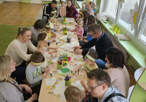 Dzieci z rodzicami siedzą wokół stołów i ozdabiają piankowe pisanki.