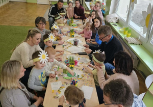 Dzieci z rodzicami siedzą wokół stołów i ozdabiają piankowe pisanki.