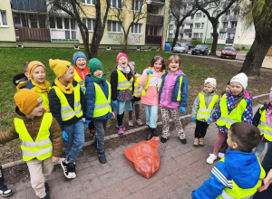 Grupa dzieci w kamizelkach stoi na chodniku. W środku stoi czerwony worek ze śmieciami.