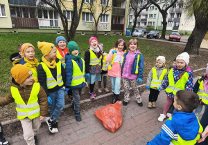 Grupa dzieci w kamizelkach stoi na chodniku. W środku stoi czerwony worek ze śmieciami.