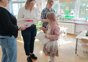 Pani Dyrektor wręcza dziewczynce nagrodę. Po bokach stoją dwie Panie prowadzące.