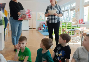 Panie prowadzące stoją z nagrodami. Na dywanie siedzą dzieci.