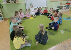 Dziewczynka siedzi na krześle w zielonej koronie. W rękach trzyma różowego balona. Wokół niej siedzą dzieci.