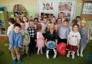 Dziewczynka siedzi w złotej koronie na krześle. Wokół niej stoją dzieci.