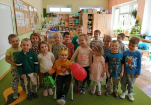 Chłopiec siedzi w koronie na krześle. W ręce trzyma balona. Wokół niego stoją dzieci.