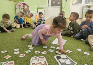 Dziewczynka w różowej bluzce i fioletowej spódniczce siedzi na zielonym dywanie i segreguje odpady.