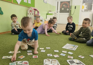 Chłopiec w niebieskiej bluzce z krótkim rękawkiem siedzi na zielonym dywanie i segreguje odpady.