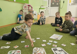Chłopiec w bluzce w zwierzątka siedzi na zielonym dywanie i segreguje odpady.