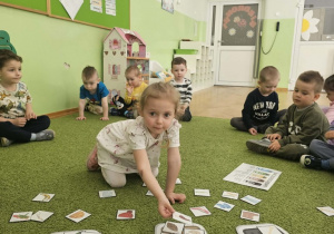 Dziewczynka w białej sukience siedzi na zielonym dywanie i segreguje odpady.