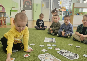 Chłopiec w żółtej bluzce siedzi na zielonym dywanie i segreguje odpady.