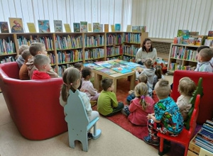 Dzieci siedzą między regałami na dywanie, na krzesełkach oraz na kanapach.