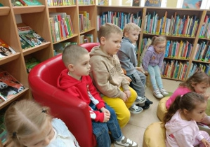 Dzieci siedzą na czerwonej kanapie. W tle widać regały z książkami.