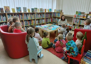 Dzieci siedzą między regałami na dywanie, na krzesełkach oraz na kanapach.
