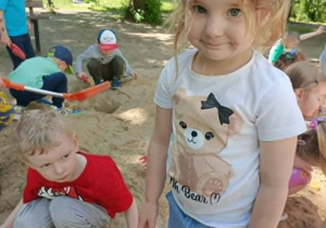 Dziewczynka w bluzce w misie pozuje do zdjęcia. W tle widać bawiące się dzieci.