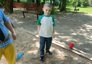 Chłopiec w biało-zielonej bluzce stoi w piaskownicy i uśmiecha się do zdjęcia.