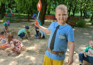 Chłopiec w niebieskiej bluzce stoi w piaskownicy, w ręce trzyma pomarańczową łopatkę i uśmiecha się do zdjęcia.