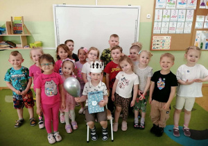Chłopiec w białej koronie i ze srebrnym balonem siedzi na krześle. Wokół stoją dzieci.