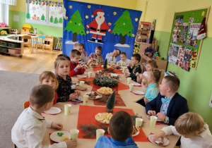 Dzieci siedzą przy wigilijnym stole w tle dekoracja z choinkami i Mikołajem