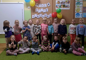 Grupa dzieci uśmiecha się do zdjęcia w tle balony i napis dzień przedszkolaka