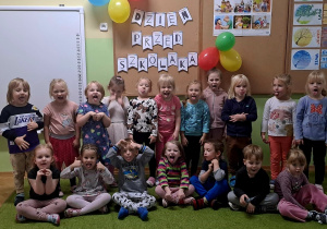 Dzieci pozują do zdjęcia z zabawnymi minami, w tle balony i tablica z napisem dzień przedszkolaka