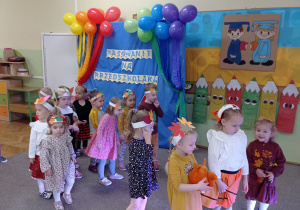 Grupa dzieci stoi na dywanie, w tle dekoracja pasowanie na przedszkolaka i balony