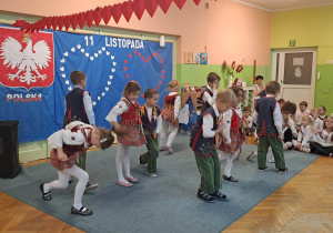 Dzieci w strojach krakowskich podczas pokazu tańca. W tle dekoracja patriotyczna