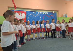 Dzieci ubrane na galowo, stoją w rzędzie i śpiewają piosenkę. W tle dekoracja patrotyczna.