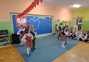 Dzieci w strojach krakowskich podczas pokazu tańca. W tle dekoracja patriotyczna