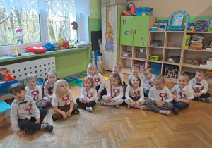 Dzieci siedzą na podłodze, na piersi mają kotylion w barwach narodowych. W tle szafki, zabawki.
