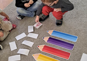 Dzieci siedzą na dywanie, chłopiec odkrywa obrazek, na podłodze leżą kolorowe sylwety kredek