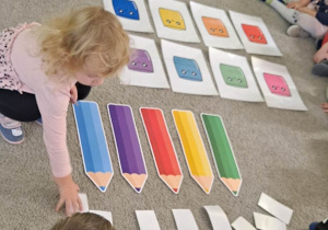 Dzieci siedzą na dywanie, dziewczynka odkrywa obrazek, na podłodze leżą kolorowe sylwety kredek