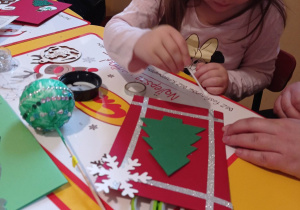 Dziewczynka siedzi przy stoliku i dekoruje kartkę świąteczną